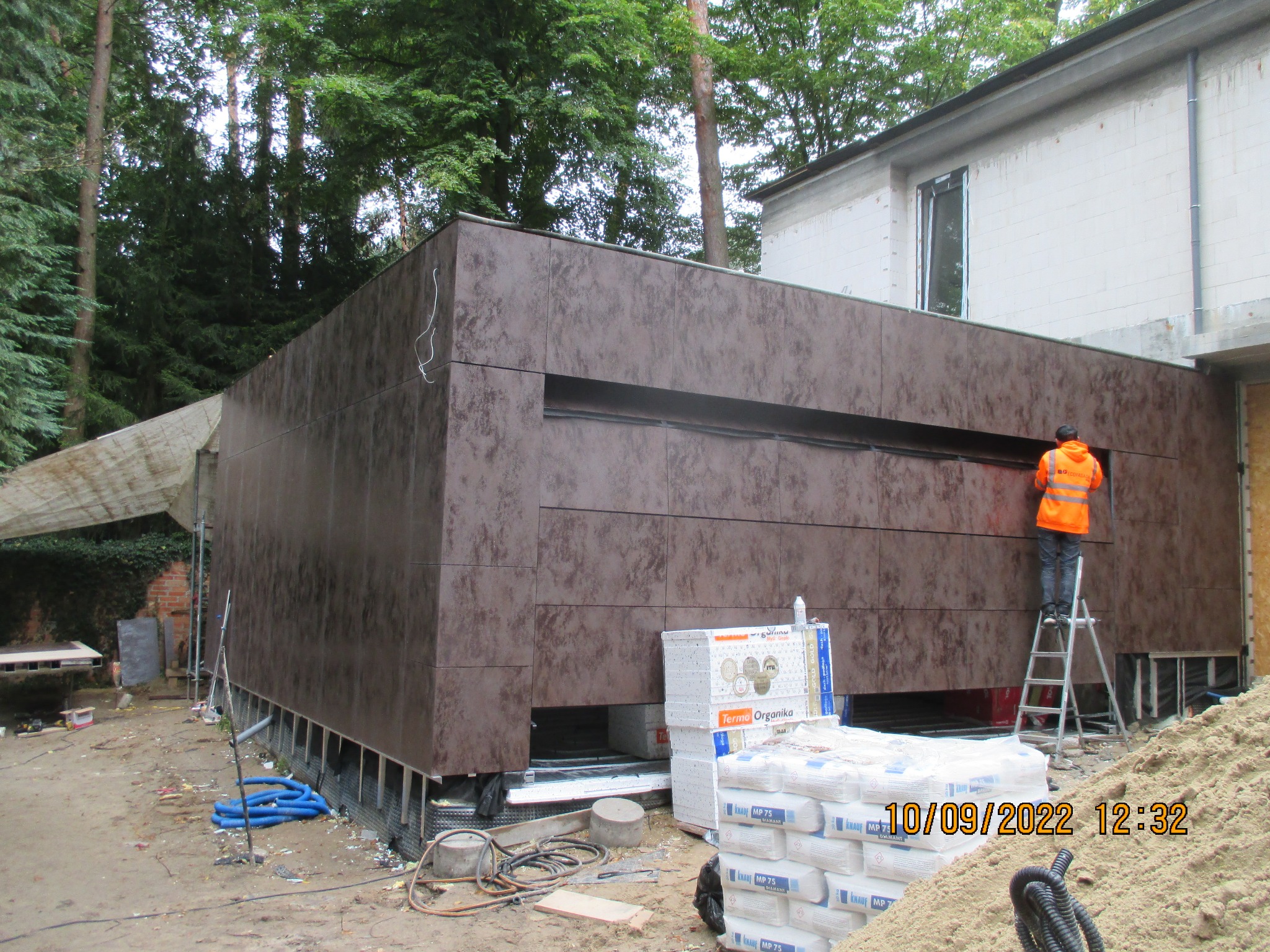 Montaż brązowej elewacji z paneli na nowym budynku, pracownik w pomarańczowej kamizelce na drabinie, widoczne materiały budowlane i elementy konstrukcyjne.