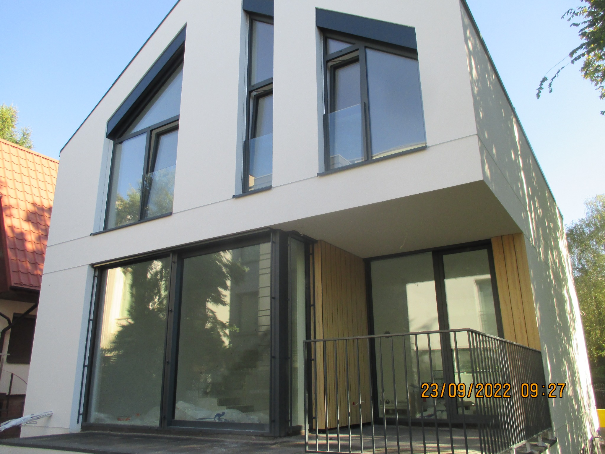 Nowoczesny dom z białą elewacją, dużymi oknami z czarnymi ramami i drewnianymi elementami wykończeniowymi, widok z dołu na fragment balkonu z metalową balustradą.