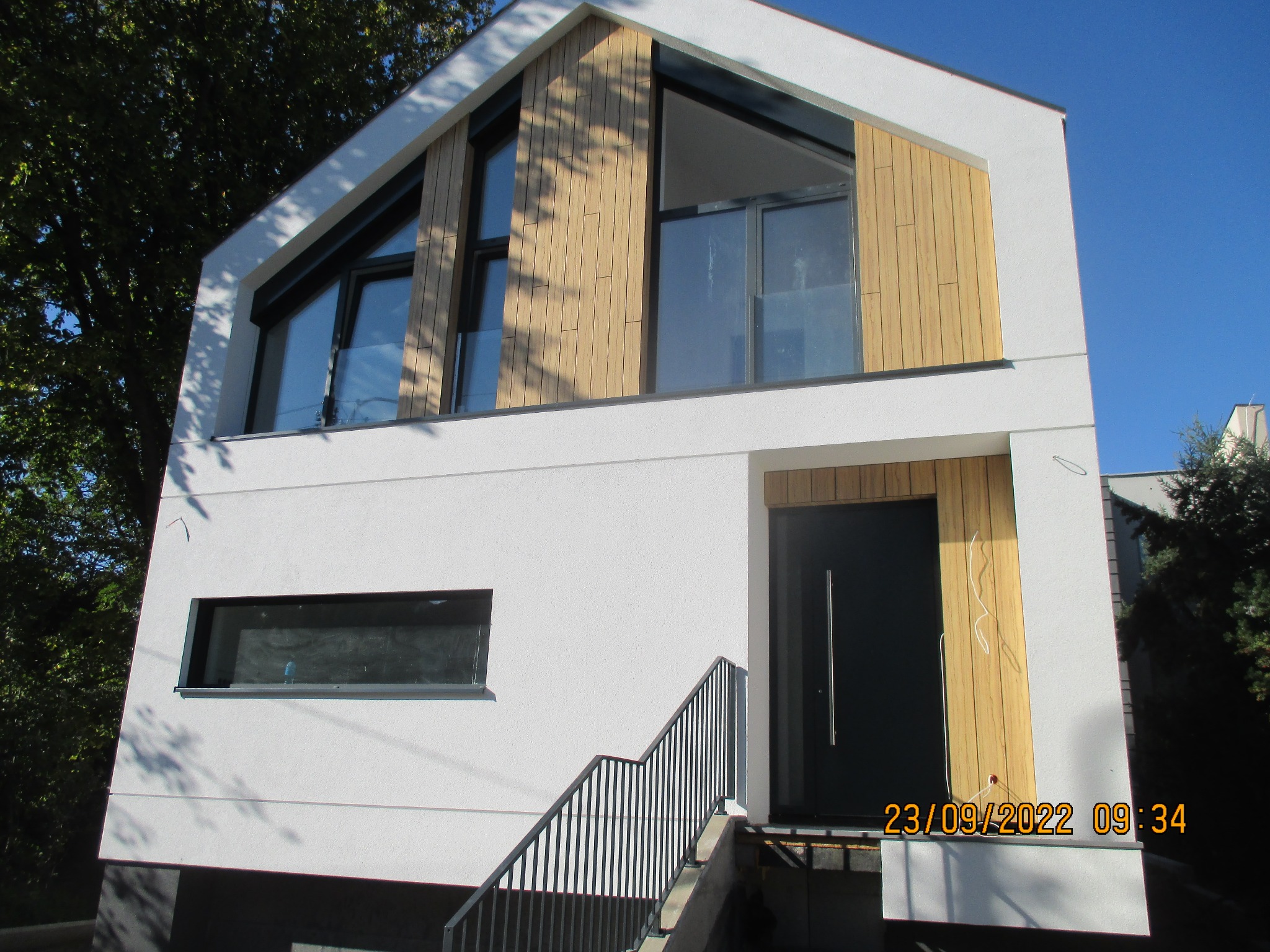 Nowoczesny dom jednorodzinny z białą elewacją i elementami drewnianymi na szczycie, widoczne schody wejściowe z czarną balustradą i czarne okna.