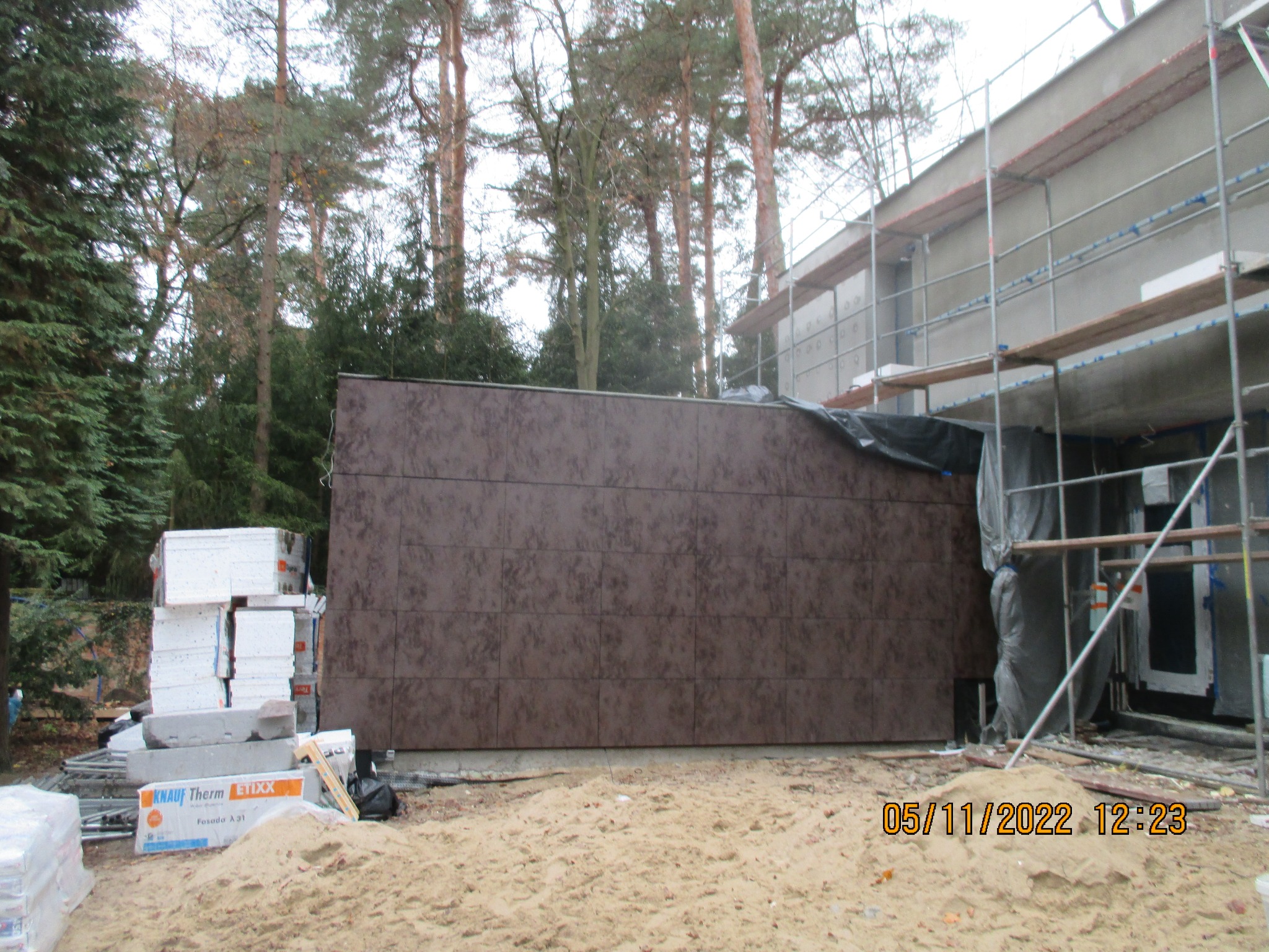 Budynek w trakcie prac elewacyjnych: widoczne rusztowania, brązowe panele elewacyjne, ocieplenie styropianowe i piasek na placu budowy, z drzewami w tle.