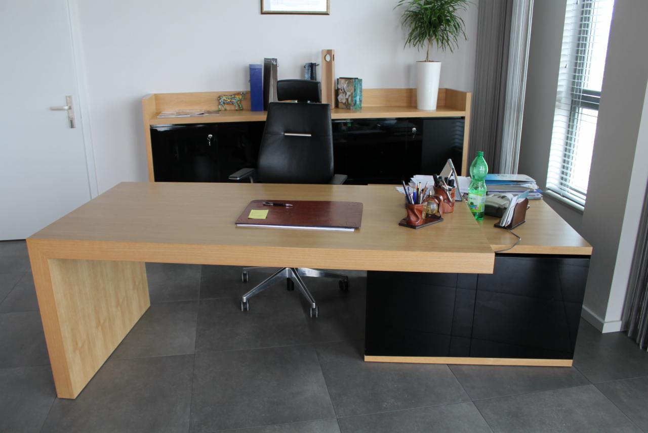 Nowoczesne biuro z drewnianym biurkiem o jasnym blacie i czarnych frontach szafek, krzesłem obrotowym, elementami dekoracyjnymi, rośliną w doniczce i zasłonami w tle.