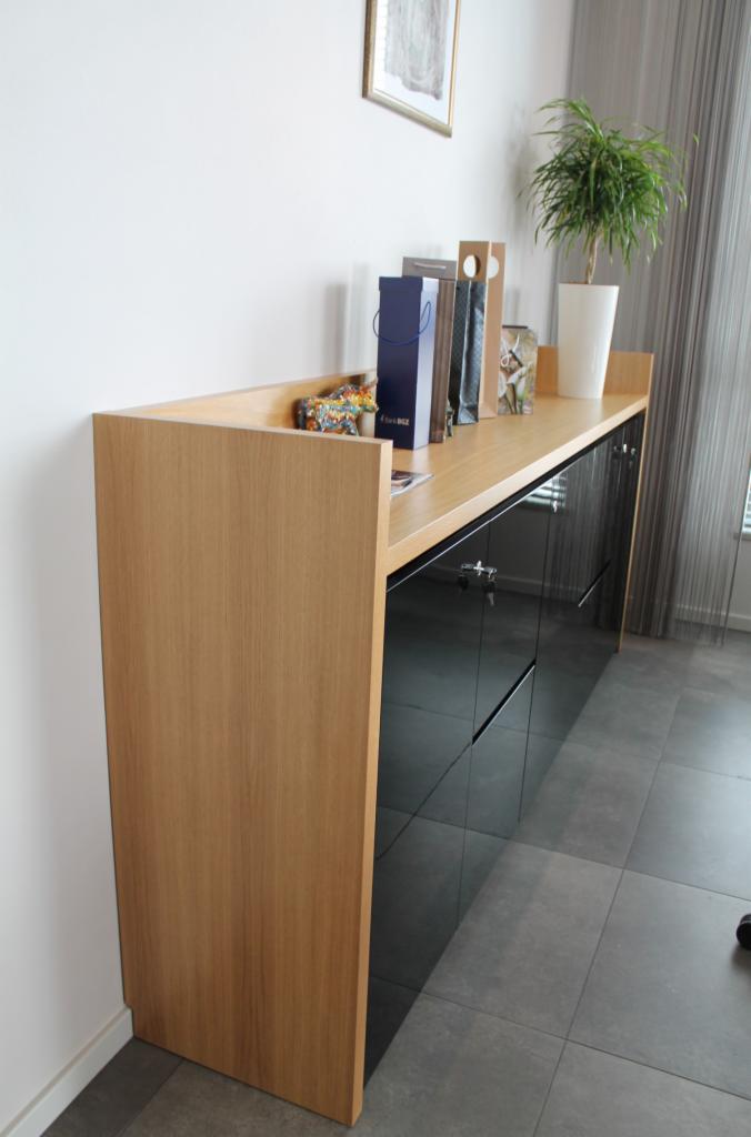 Nowoczesna komoda z drewnianym blatem i czarnymi, lakierowanymi frontami szuflad, na której stoją dekoracyjne przedmioty i doniczka z zieloną rośliną.