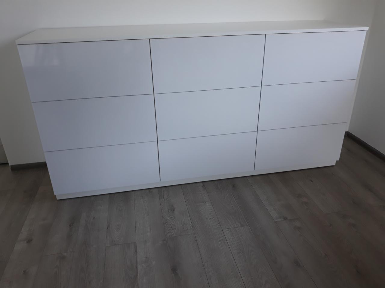 Biała, minimalistyczna szafa z dziewięcioma frontami, umieszczona na tle szarej podłogi z paneli, z widocznymi detalami łączeń i subtelnymi refleksami światła.