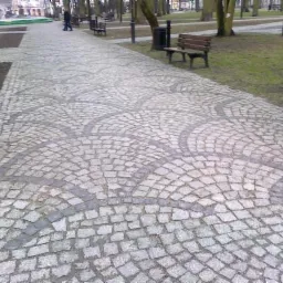 Ścieżka w parku wyłożona szarą kostką, ułożona we wzór koncentrycznych łuków z ciemniejszej kostki, w tle drzewa i ławki.