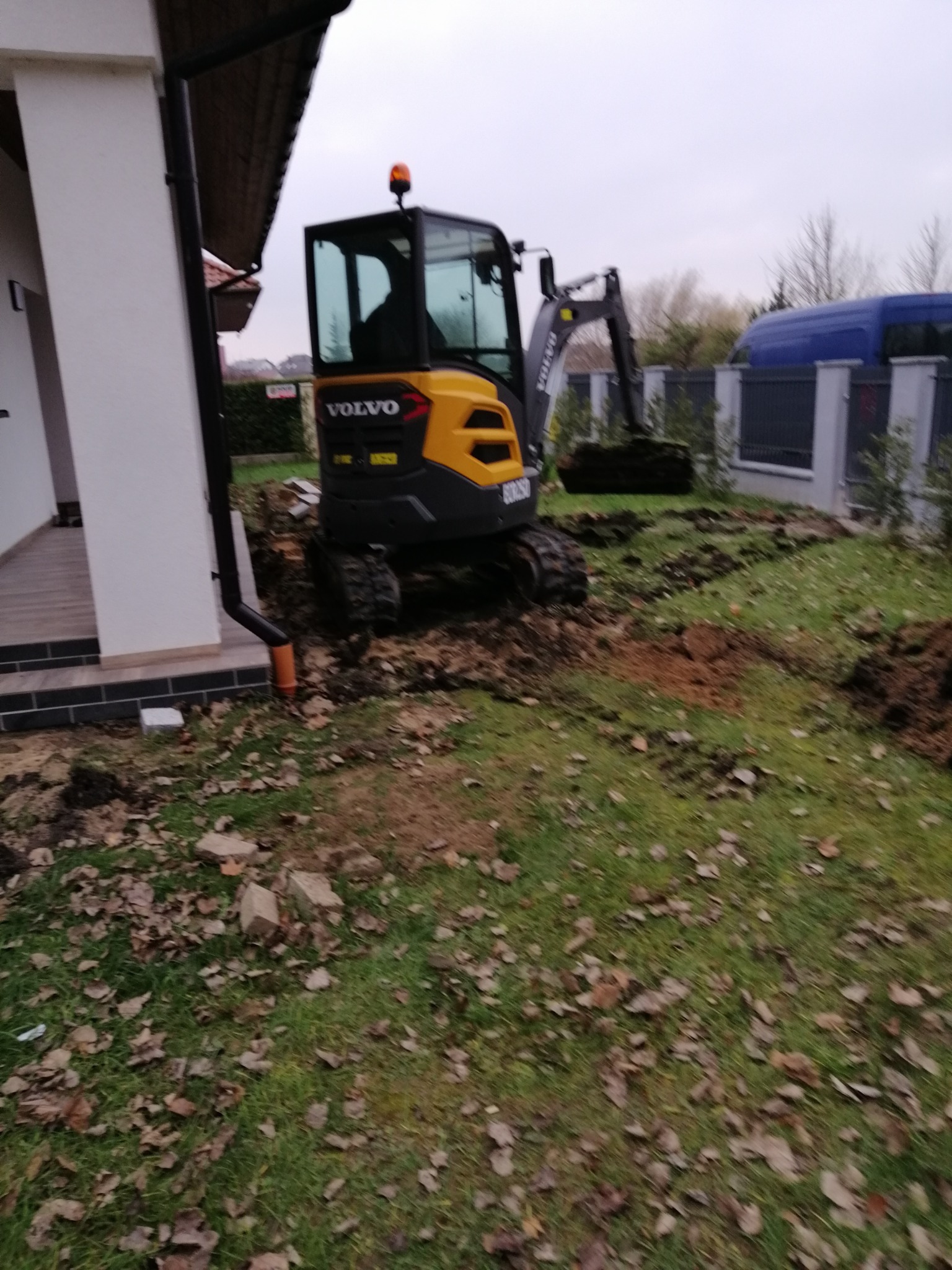 Żółto-szara minikoparka Volvo na trawniku obok domu, przygotowująca teren pod budowę lub remont, widoczna wykopana ziemia.