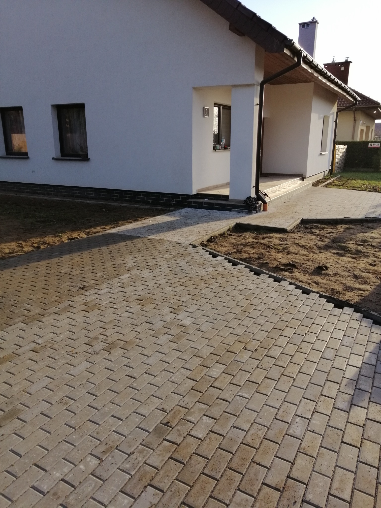 Nowo ułożona, beżowa kostka brukowa prowadząca do jasnego domu jednorodzinnego z gankiem, widoczne fragmenty przygotowanego podłoża.