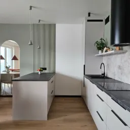 Nowoczesna kuchnia z wyspą, czarnym blatem i białymi szafkami. W tle widoczna jadalnia przez łukowe przejście. Minimalistyczne oświetlenie.