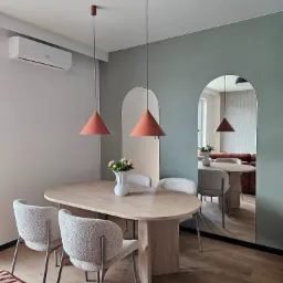 Nowoczesna jadalnia z owalnym stołem, teksturowanymi krzesłami i pomarańczowymi lampami stożkowymi. Lustra w kształcie łuków odbijają przestrzeń. Klimatyzator na ścianie.