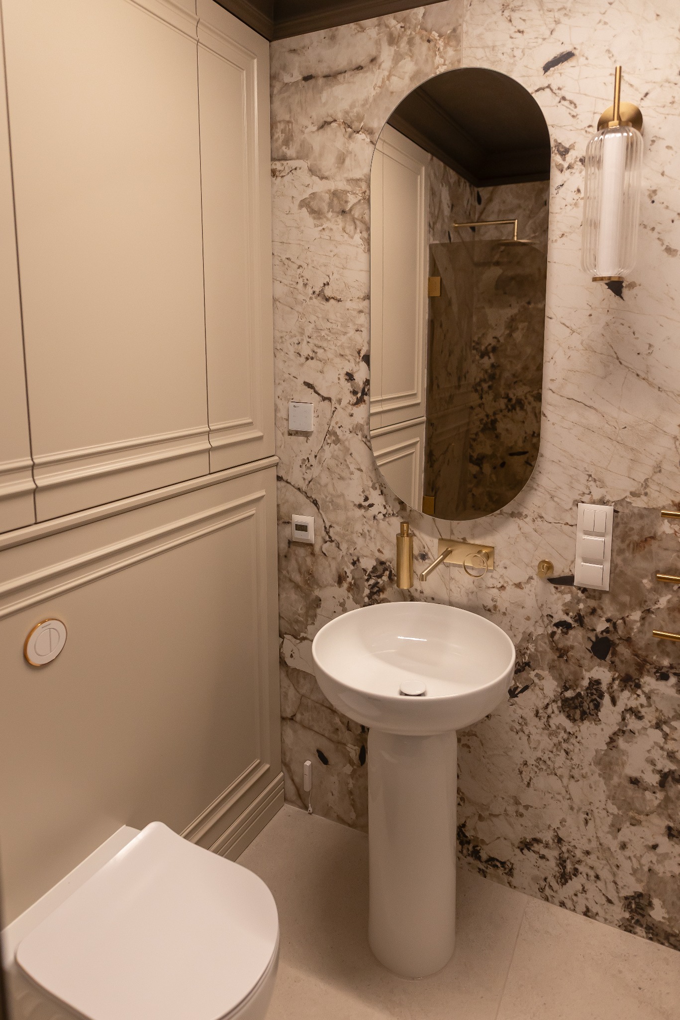 Nowoczesna łazienka z umywalką na postumencie, marmurową ścianą i owalnym lustrem z podświetleniem. Widoczne złote detale i minimalistyczna toaleta.