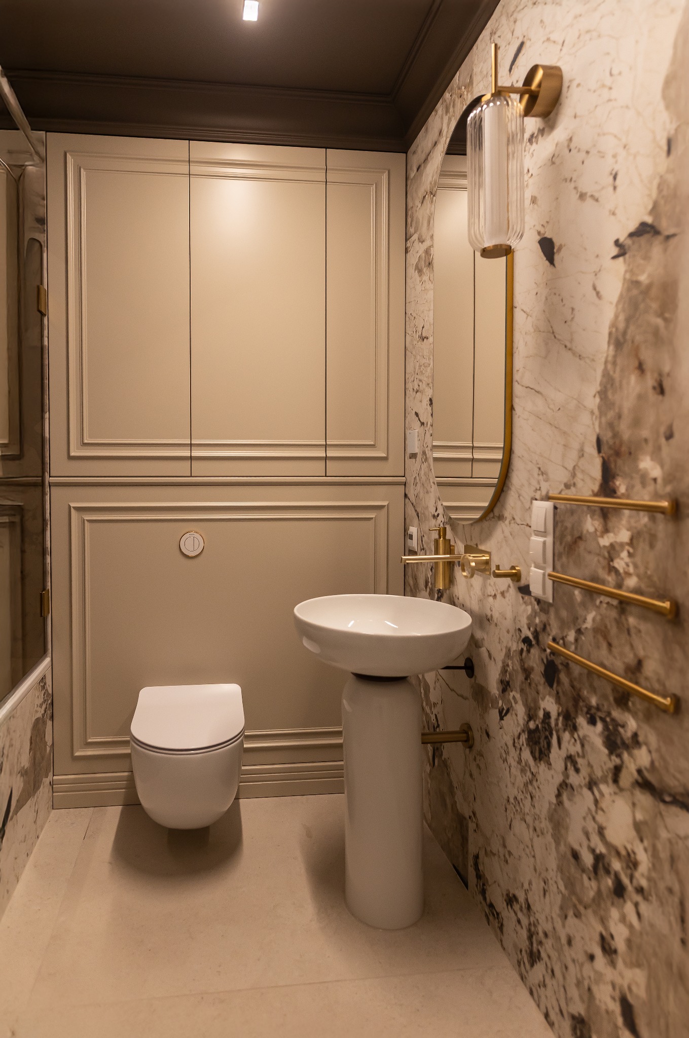 Elegancka łazienka z umywalką na postumencie, toaletą w zabudowie i marmurową ścianą. Złote akcenty: armatura, oświetlenie, grzejnik drabinkowy. Nowoczesne wykończenie.