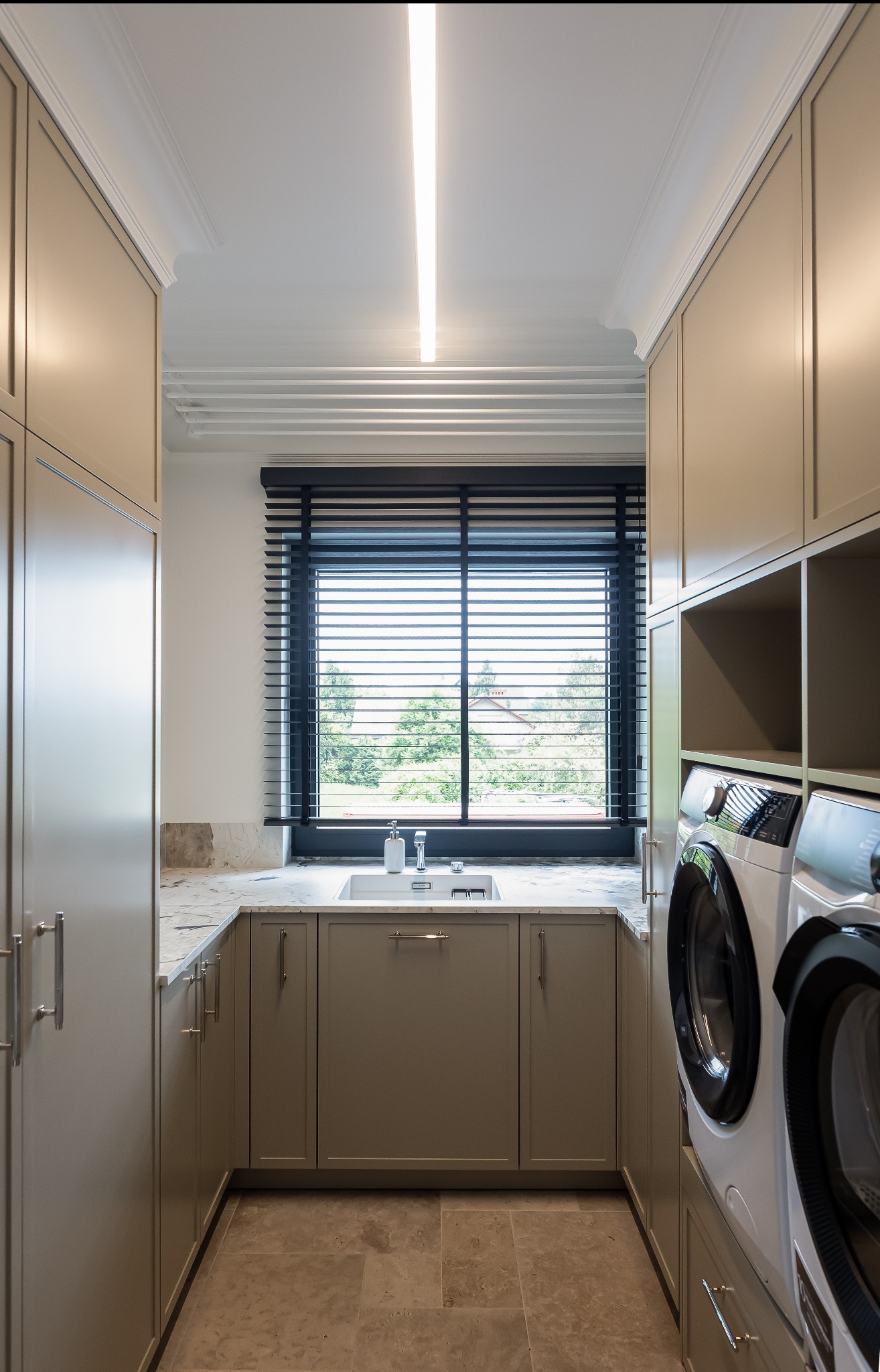 Nowoczesna pralnia w beżowych odcieniach: zabudowa meblowa, marmurowy blat z umywalką przy oknie z żaluzjami, pralka i suszarka. Minimalistyczny design, eleganckie wykończenie.