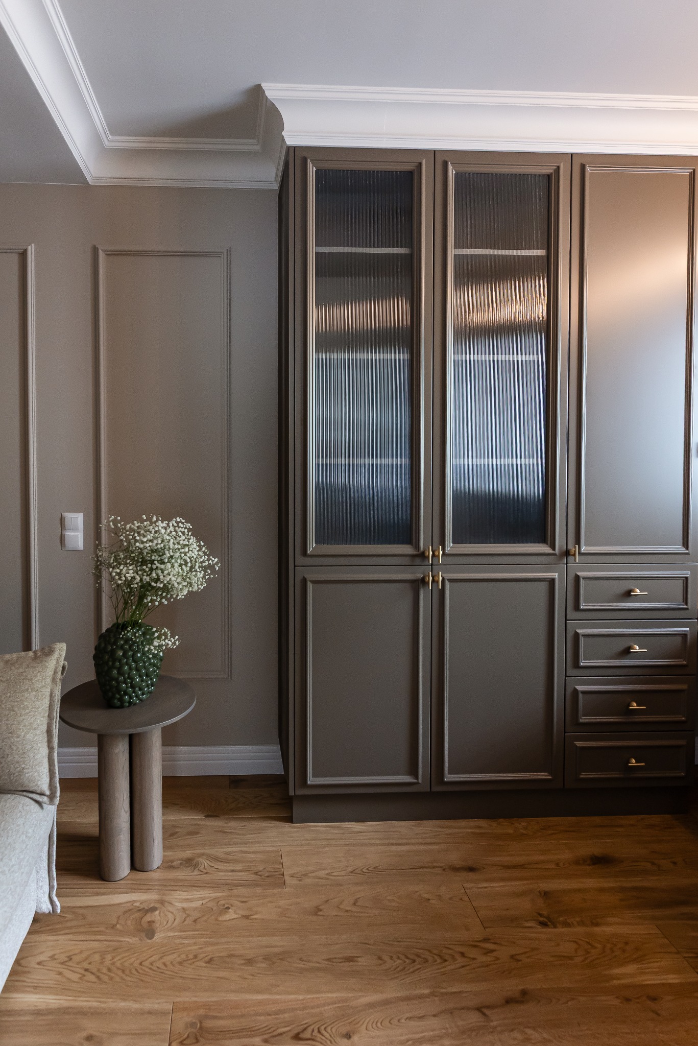 Stylowe wnętrze z drewnianą podłogą, zabudową w kolorze taupe z ryflowanymi szybami i dekoracyjną sztukaterią. Na stoliku bukiet gipsówki w okrągłym wazonie.