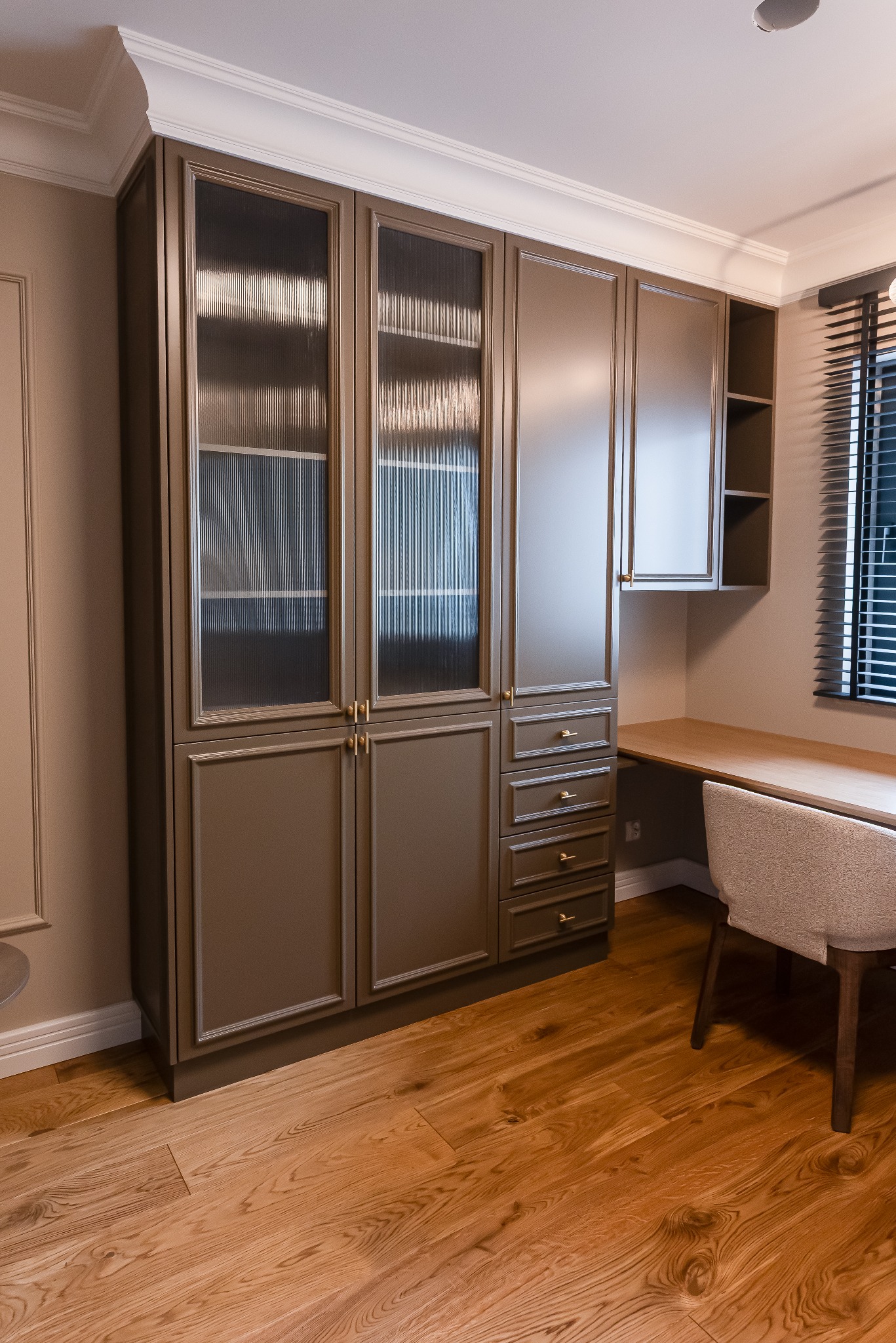 Elegancki gabinet z zabudową meblową w odcieniach brązu i beżu, z biurkiem i krzesłem. Ryflowane szkło w witrynach dodaje subtelnej tekstury. Całość dopełnia drewniana podłoga.