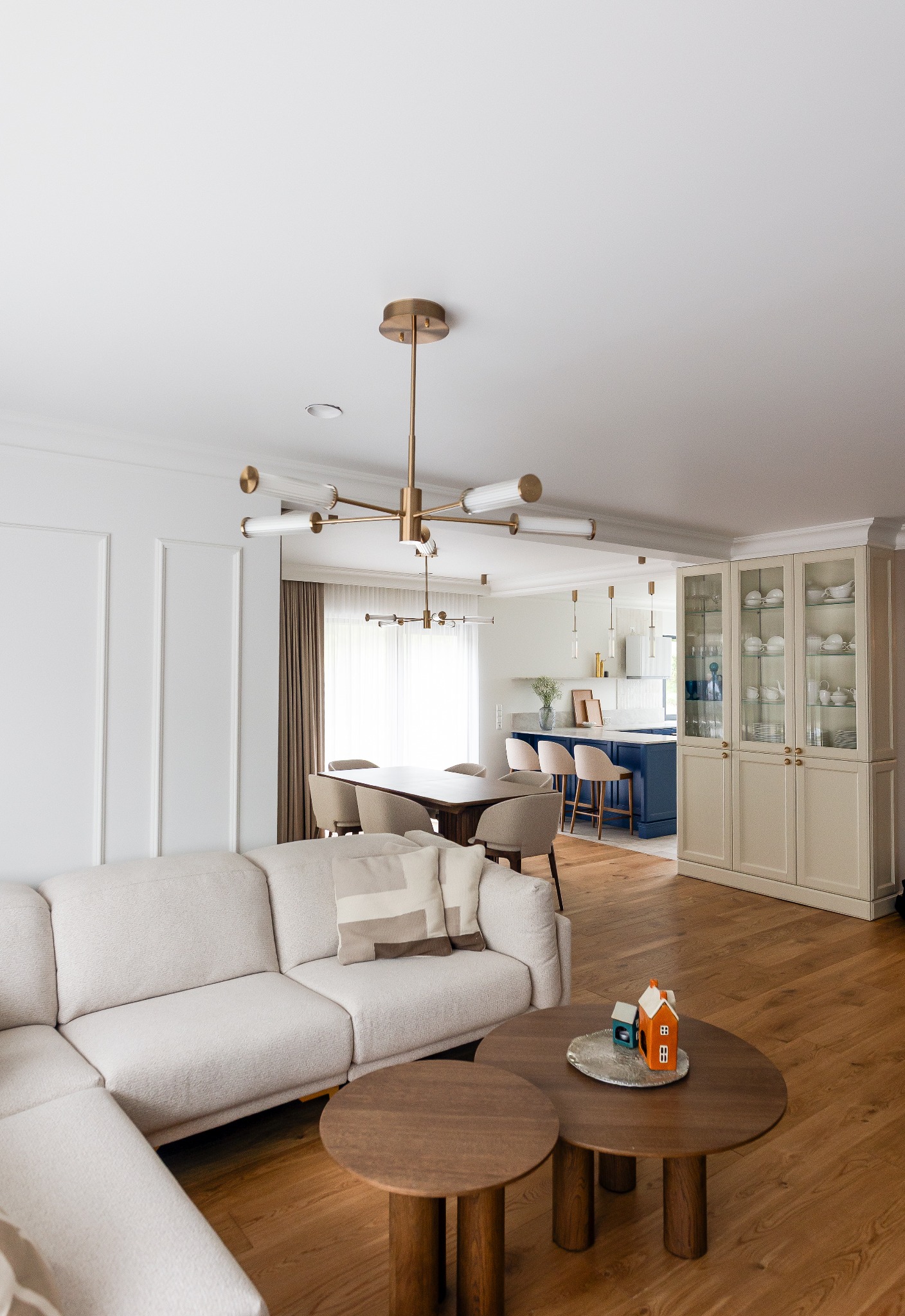 Jasny salon z drewnianą podłogą, beżową sofą i okrągłymi stolikami kawowymi. W tle jadalnia i kuchnia z niebieską wyspą, całość w nowoczesnym stylu.