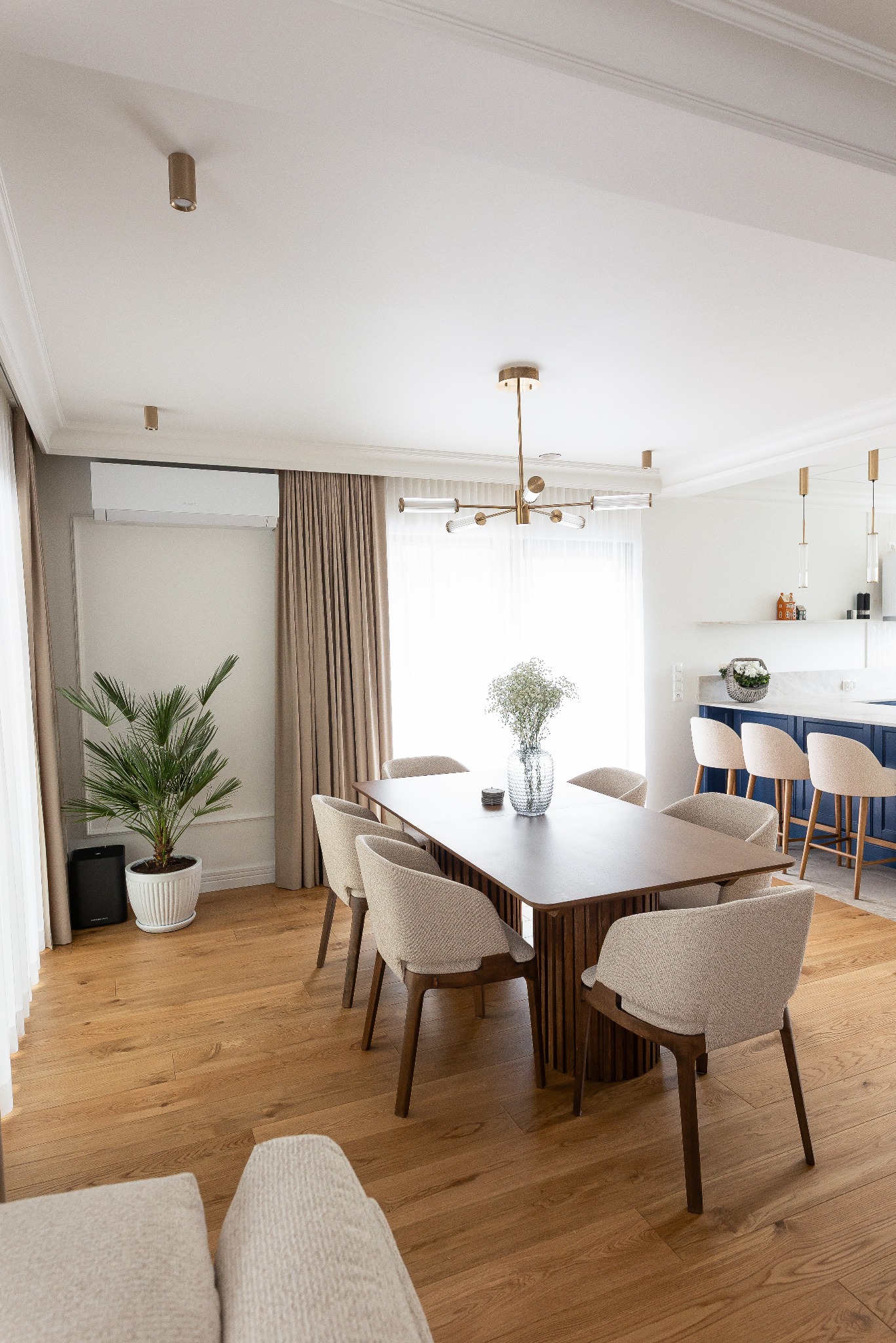Jasne, przestronne wnętrze jadalni z drewnianym stołem, krzesłami z beżową tapicerką, parkietem i rośliną doniczkową. W tle widoczna kuchnia z niebieską zabudową.