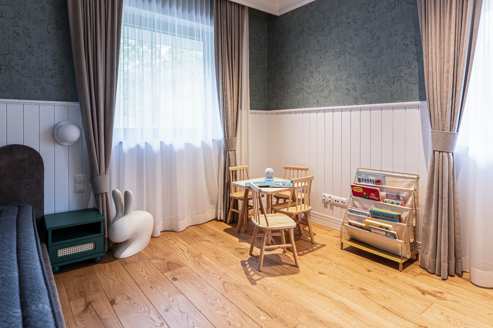 Jasny pokój dziecięcy z drewnianą podłogą, stolikiem i krzesłami, regałem na książki oraz dekoracyjnym królikiem. Ściany zdobią tapety i biała boazeria.