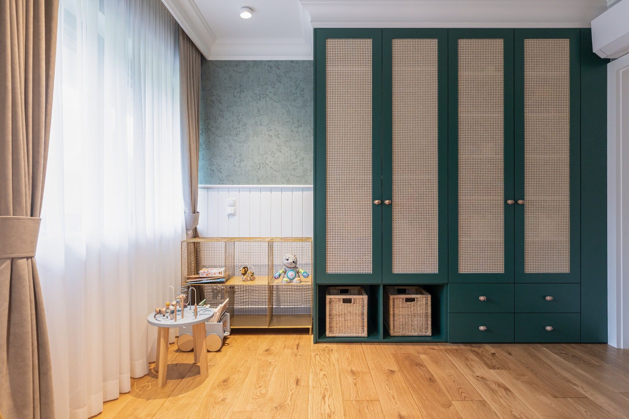 Pokój dziecięcy: zielona szafa z rattanowymi frontami, drewniana podłoga, zabawki na metalowym regale, okno z zasłonami i wzorzysta tapeta. Jasne, przestronne wnętrze.