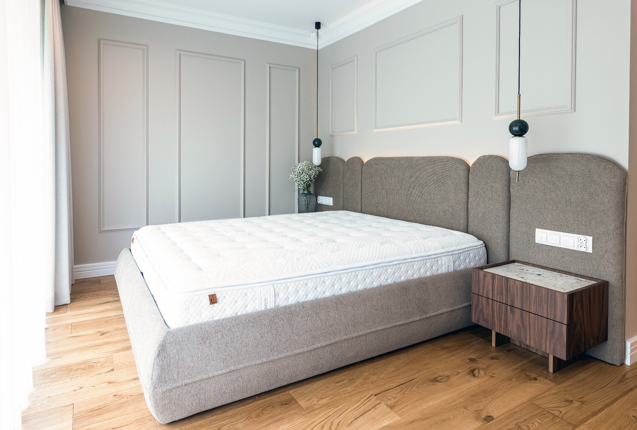 Elegancka sypialnia w stonowanych barwach: beżowe ściany z dekoracyjnymi panelami, drewniana podłoga, łóżko z tapicerowanym wezgłowiem i marmurowa szafka nocna. Minimalistyczne lampy wiszące.