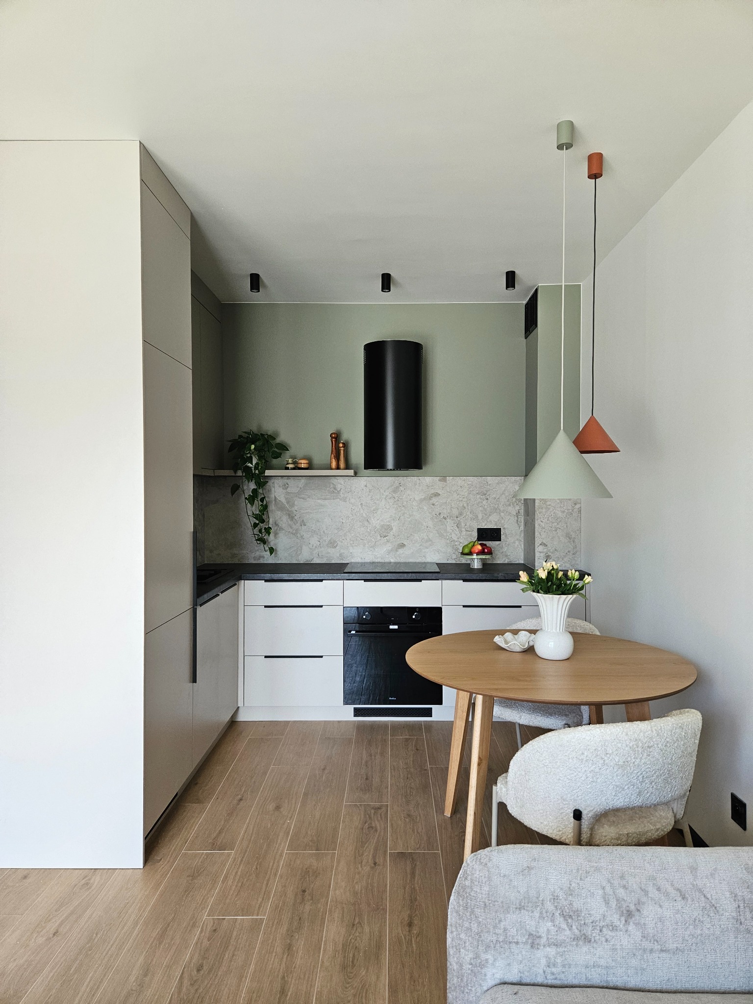 Jasna kuchnia z drewnianą podłogą i okrągłym stołem. Nad stołem wiszą dwie lampy o stożkowych abażurach w kolorach pastelowej zieleni i terakoty.