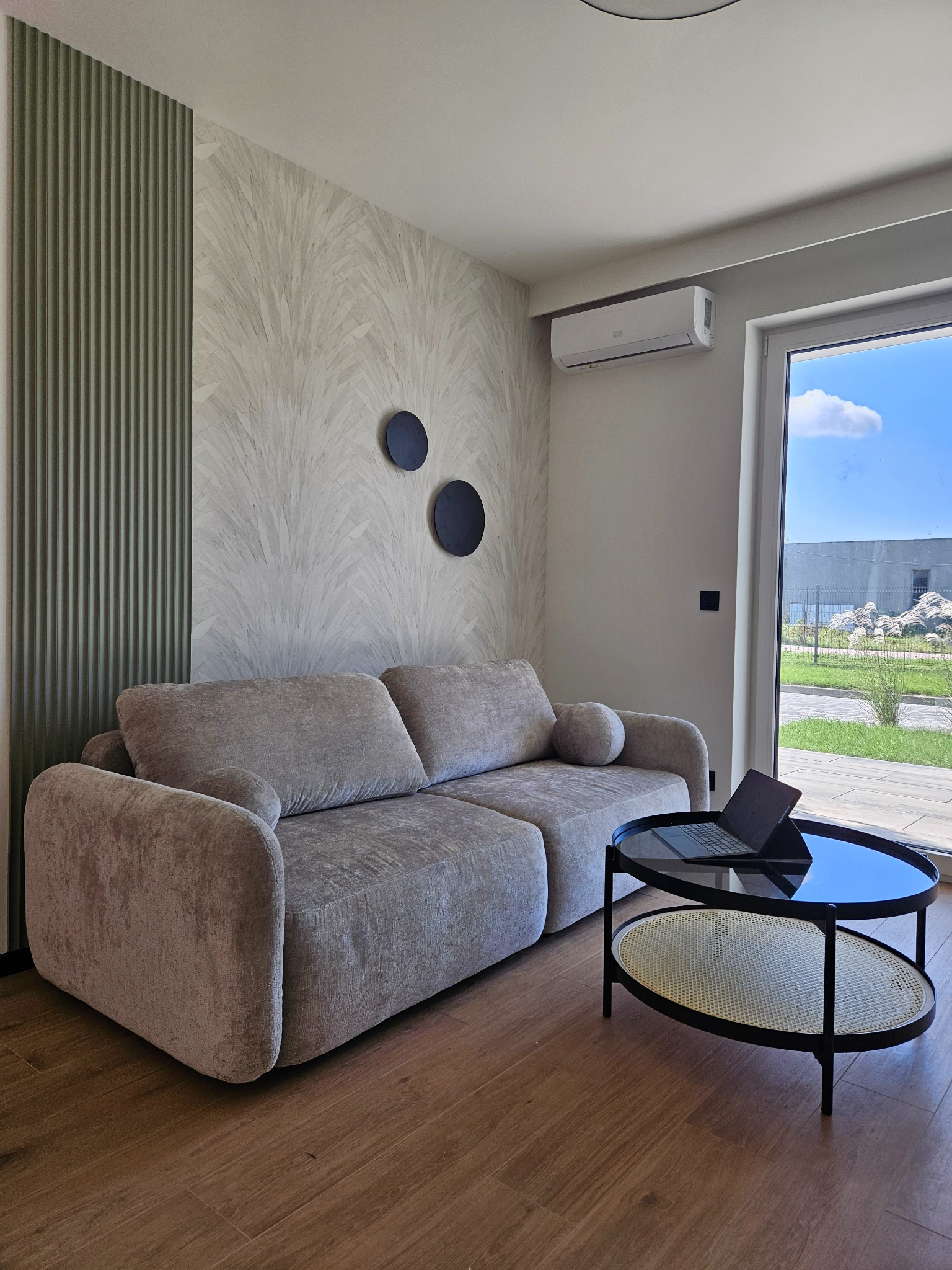 Jasny salon z szarą sofą, okrągłym stolikiem z laptopem i dekoracyjną ścianą z panelami i tapetą. Widok na zewnątrz przez duże okno.