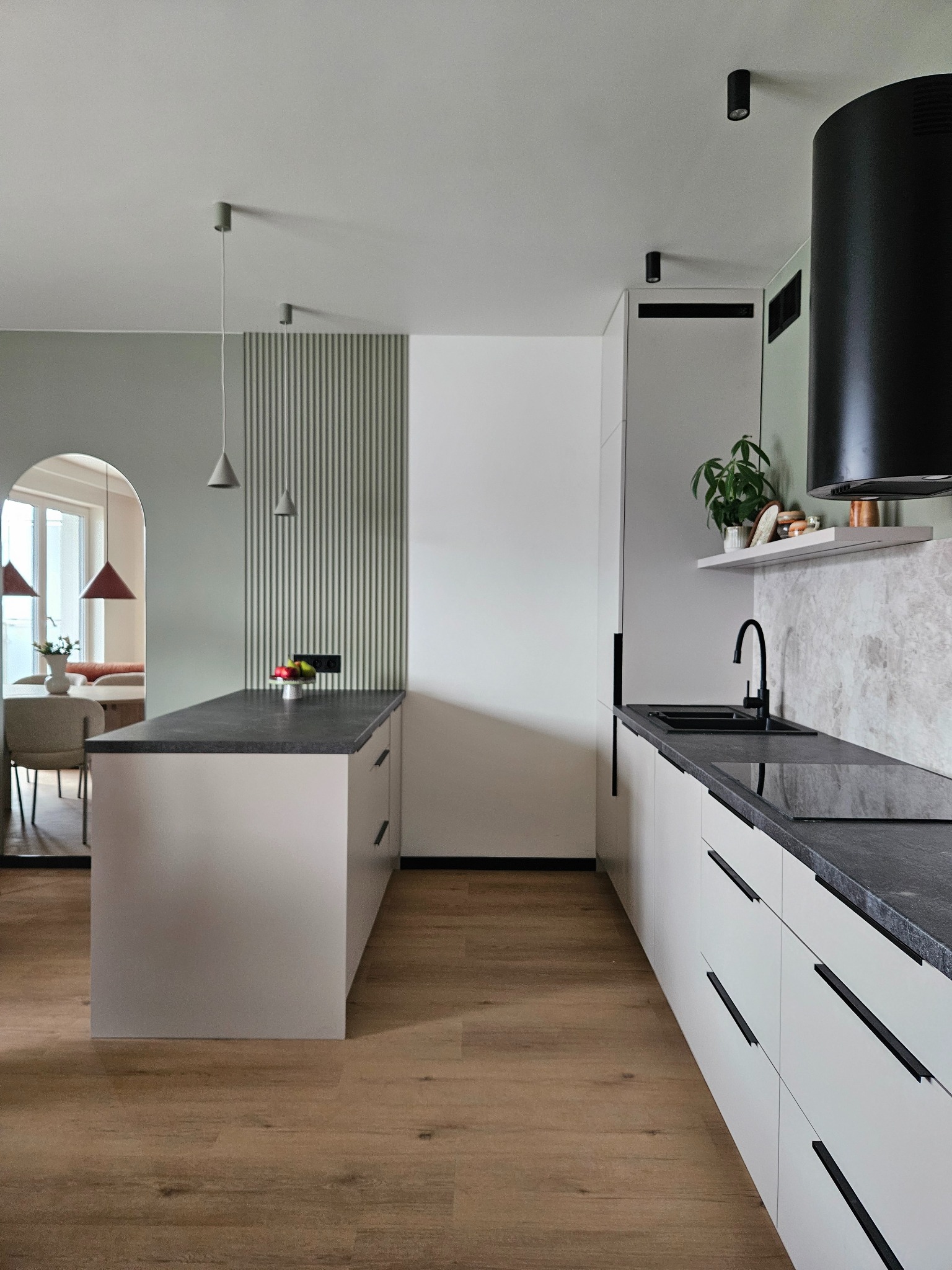 Nowoczesna kuchnia z wyspą, czarnym blatem i białymi szafkami. W tle widoczna jadalnia przez łukowe przejście. Minimalistyczne oświetlenie.