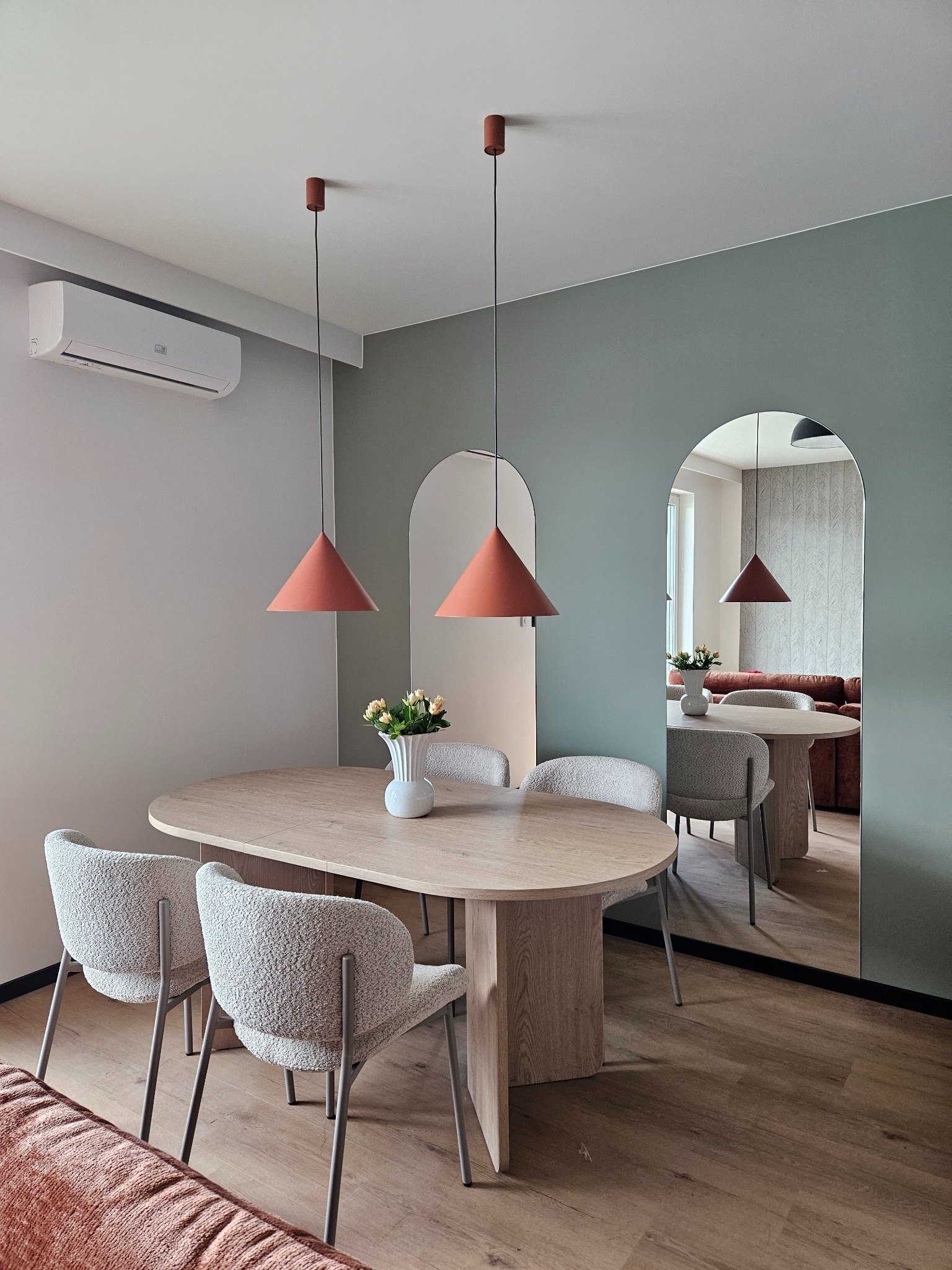 Nowoczesna jadalnia z owalnym stołem, teksturowanymi krzesłami i pomarańczowymi lampami stożkowymi. Lustra w kształcie łuków odbijają przestrzeń. Klimatyzator na ścianie.