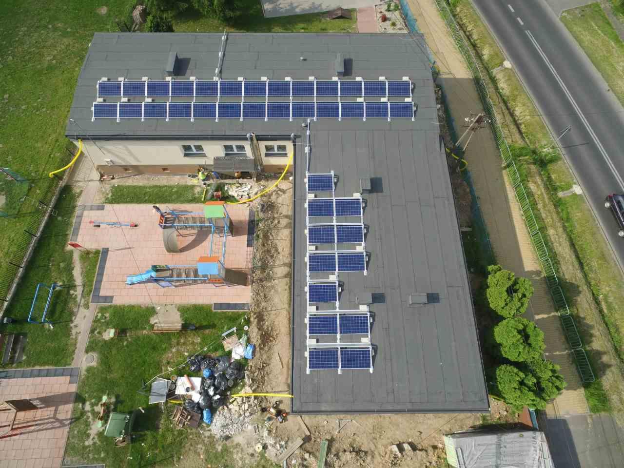 Widok z góry na budynek z panelami słonecznymi na dachu, obok plac zabaw i droga. Żółte rury prowadzą od paneli. Częściowo widoczny plac budowy.