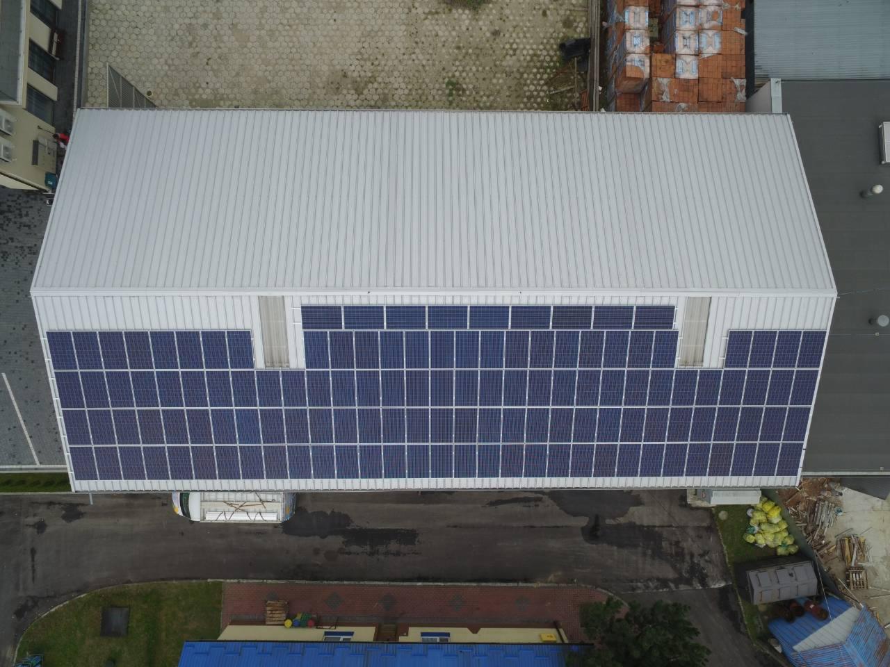 Widok z góry na duży budynek z panelami słonecznymi na ścianie, obok zaparkowany biały bus i składowane materiały budowlane. Dach blaszany, trapezowy.