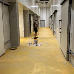 Malowanie posadzki w chłodni przemysłowej na kolor żółty, widoczni dwaj pracownicy z wałkami malarskimi, wiadro z farbą i zabezpieczony odpływ podłogowy.