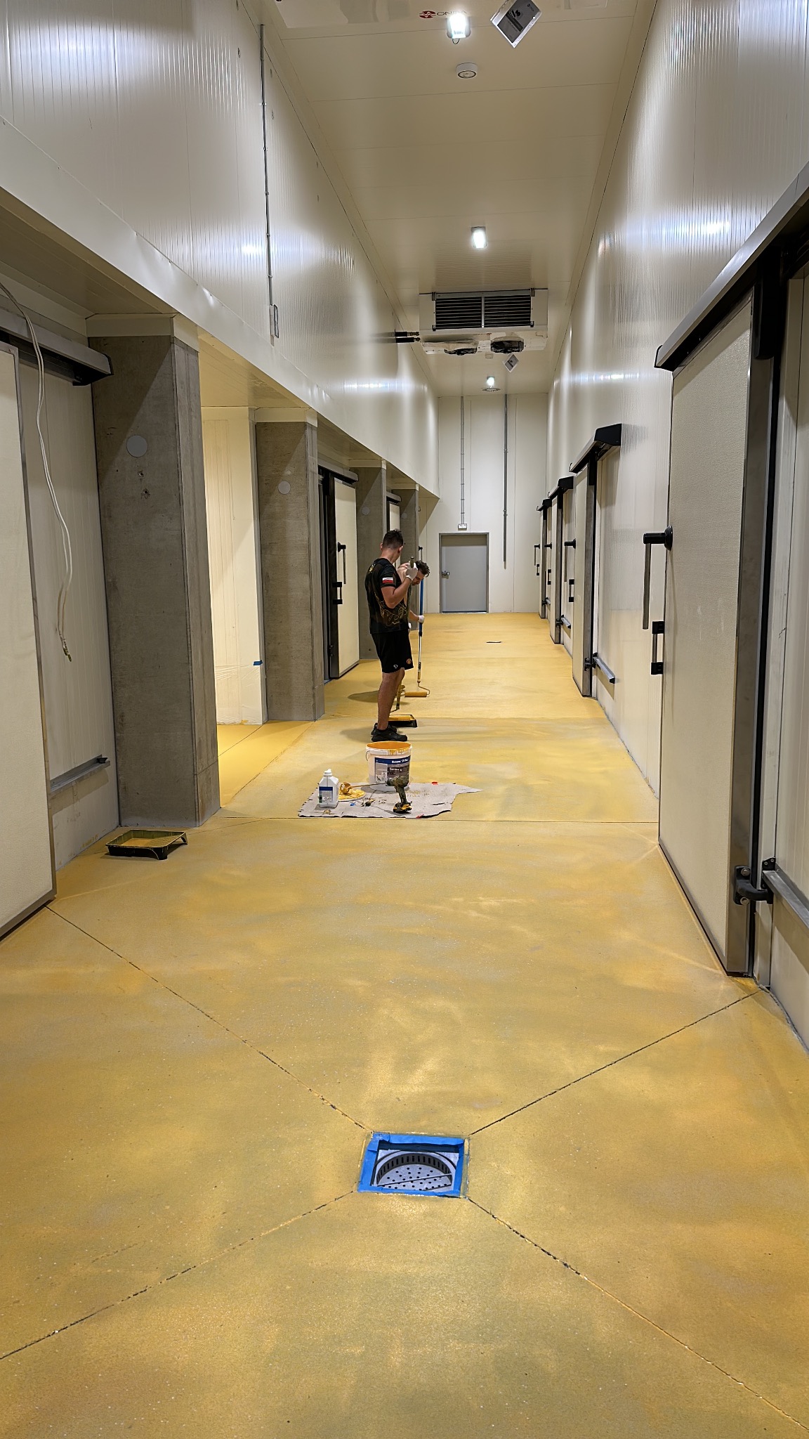 Malowanie posadzki w chłodni przemysłowej na kolor żółty, widoczni dwaj pracownicy z wałkami malarskimi, wiadro z farbą i zabezpieczony odpływ podłogowy.