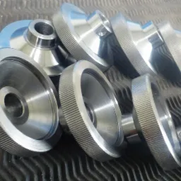 Srebrne, metalowe elementy obrotowe z radełkowaną powierzchnią na ciemnym, pofalowanym tle, widoczne otwory i precyzyjne wykończenie.