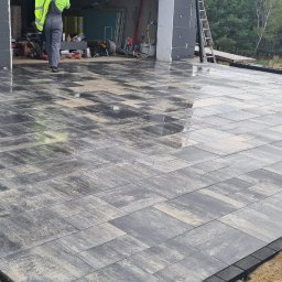 Damian - Częściowo ułożona, wilgotna kostka brukowa w odcieniach szarości i beżu, z widocznym pracownikiem w odblaskowej kamizelce i garażem w tle, na etapie wykończenia.