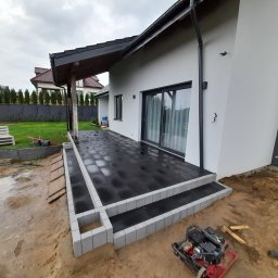 Damian - Nowoczesny taras z czarnej kostki brukowej z szarymi obrzeżami, widoczny w trakcie budowy, z ubijarką mechaniczną i pracownikiem w tle.