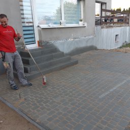 Damian - Pracownik zamiata piasek ze świeżo ułożonej, szarej kostki brukowej na podjeździe przed domem z widocznymi schodami wejściowymi i oknem.