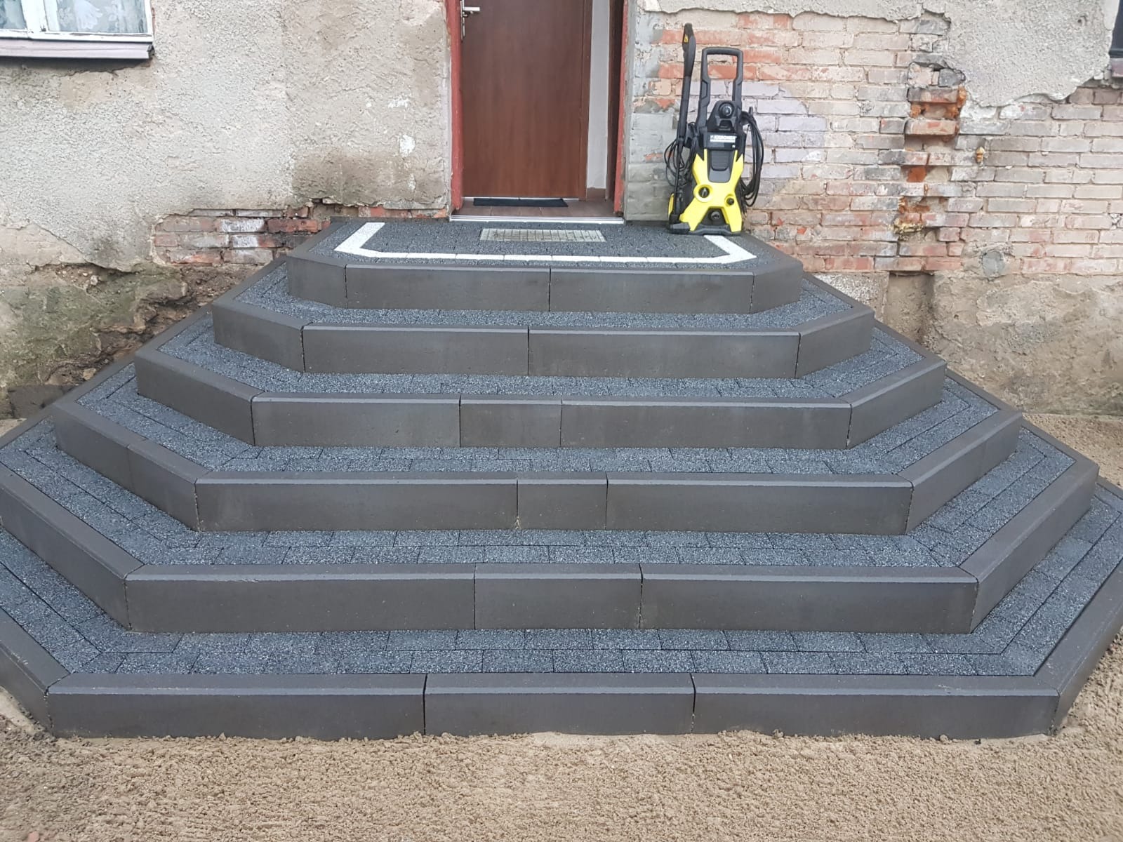 Schody wejściowe wyłożone szarą kostką brukową, z widoczną myjką ciśnieniową Karcher na górnym stopniu, na tle starego budynku z ceglaną ścianą.
