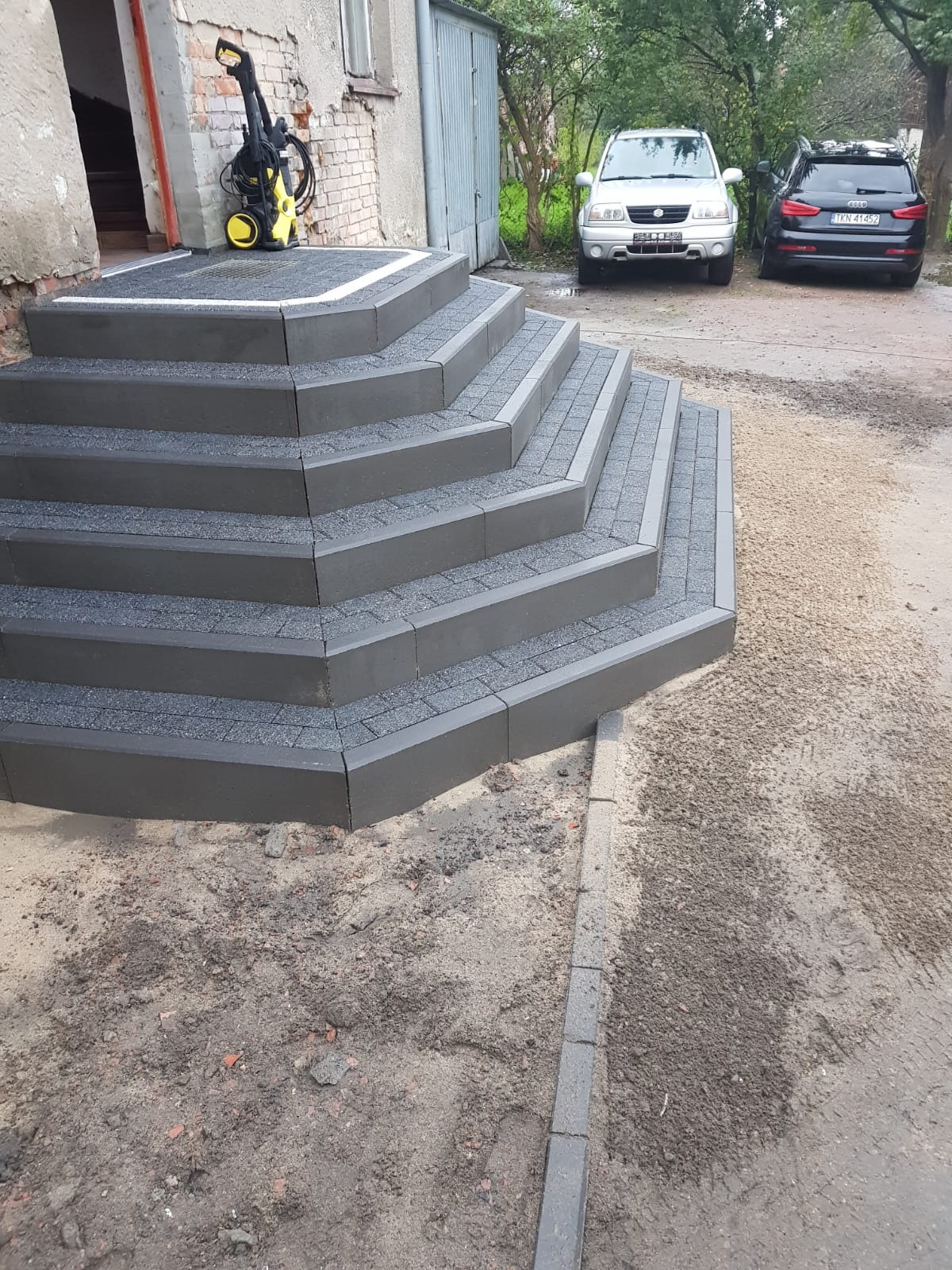 Schody wejściowe wyłożone szarą kostką brukową, z widoczną myjką ciśnieniową i dwoma samochodami w tle.