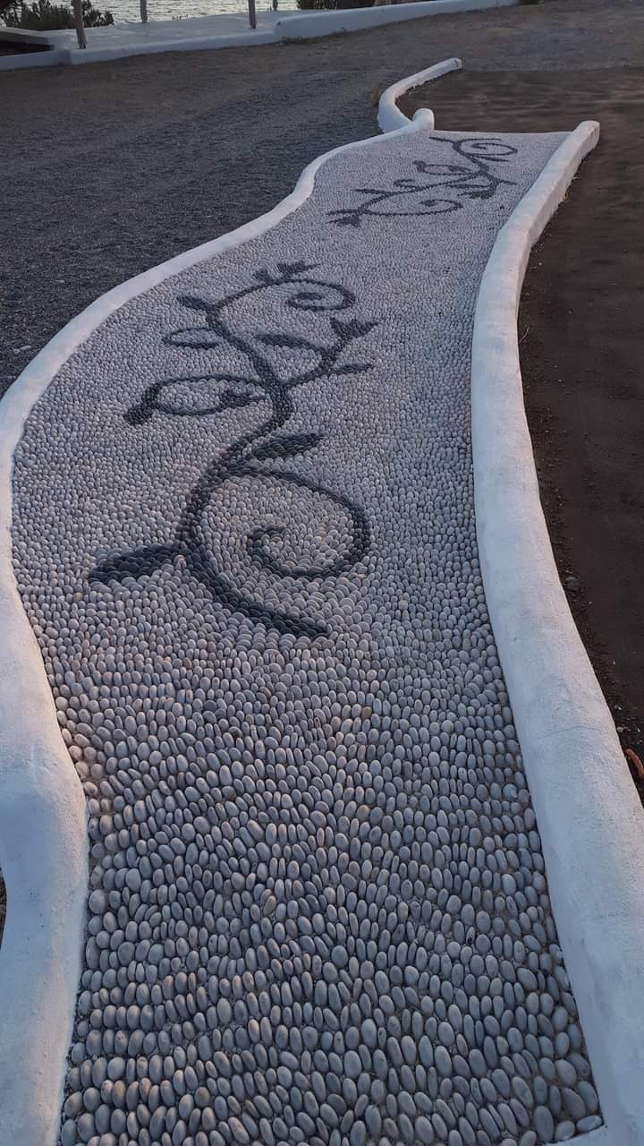 Ścieżka z jasnych, okrągłych kamieni z ciemnymi kamieniami ułożonymi w motywy roślinne, obramowana białym betonowym krawężnikiem.