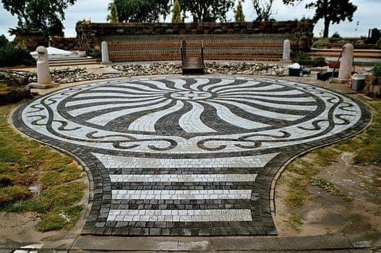 Okrągły plac z kostki brukowej ułożonej we wzór promieni słońca, z centralnym zegarem słonecznym, otoczony zielenią i elementami małej architektury.