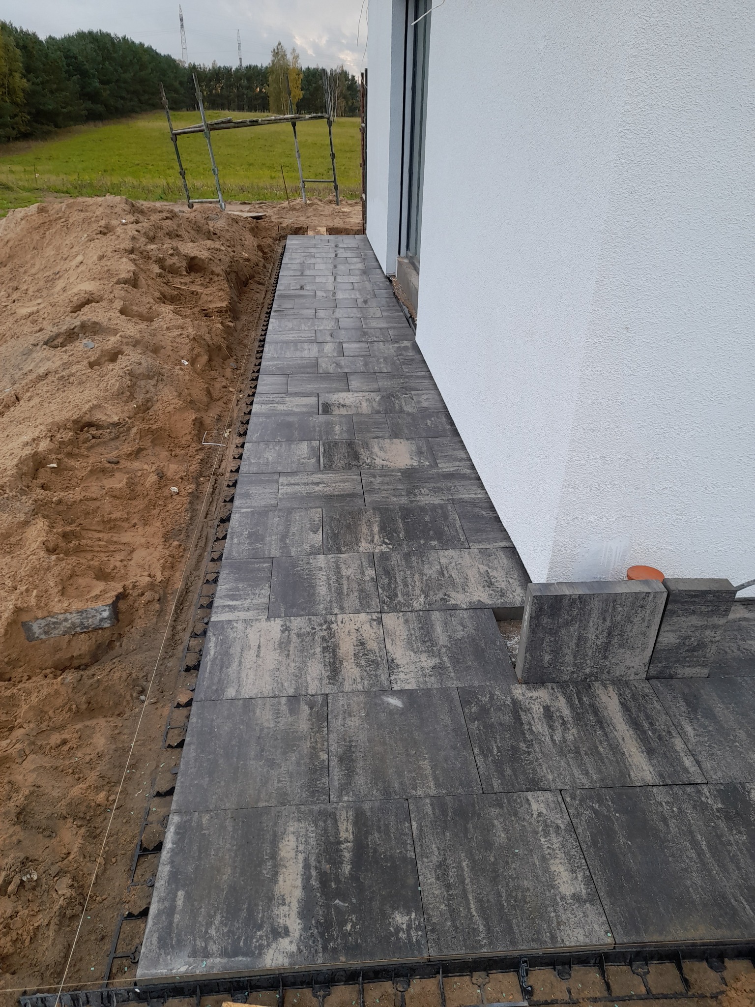 Kładzenie szarej kostki brukowej przy białym budynku, widoczne niedokończone obrzeże, ziemia i rusztowanie w tle.