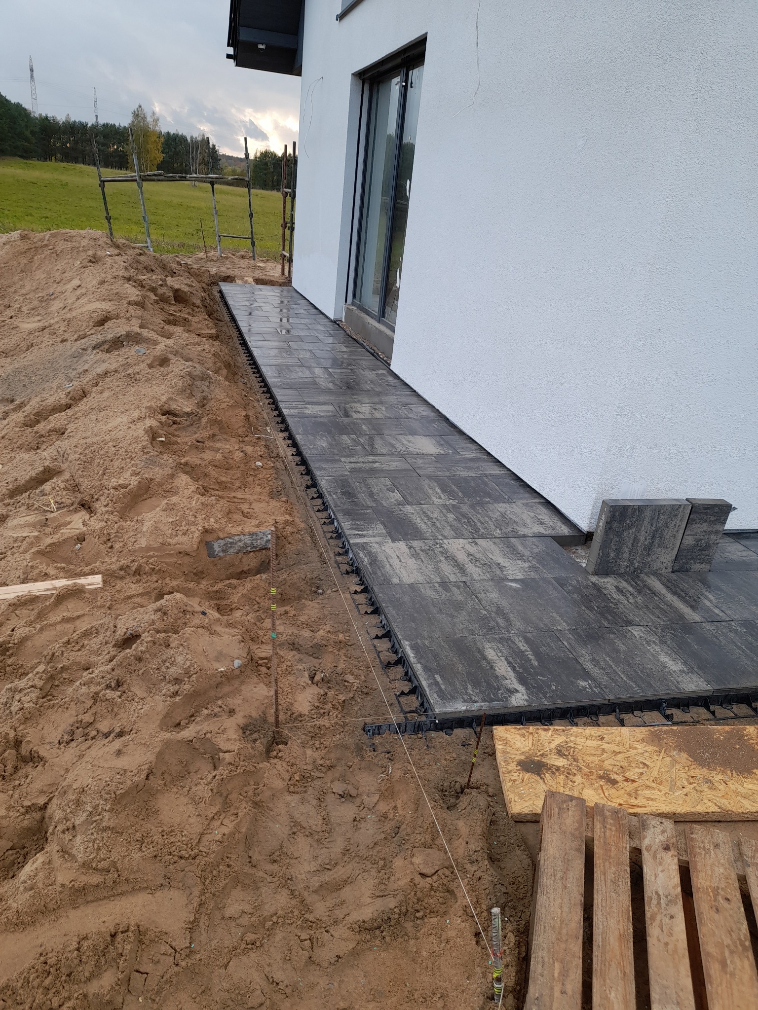 Ciemnoszara kostka ułożona przy białym budynku, widoczne elementy mocujące kostkę i przygotowane podłoże z piasku.