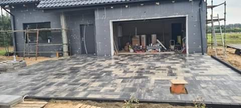 Układanie kostki brukowej na podjeździe do domu w trakcie budowy, widoczne rusztowania i materiały budowlane w otwartym garażu.