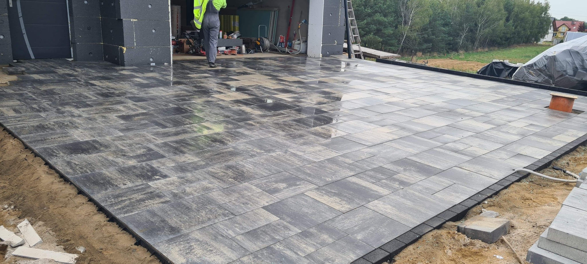 Częściowo ułożona, wilgotna kostka brukowa w odcieniach szarości i beżu, z widocznym pracownikiem w odblaskowej kamizelce i garażem w tle, na etapie wykończenia.