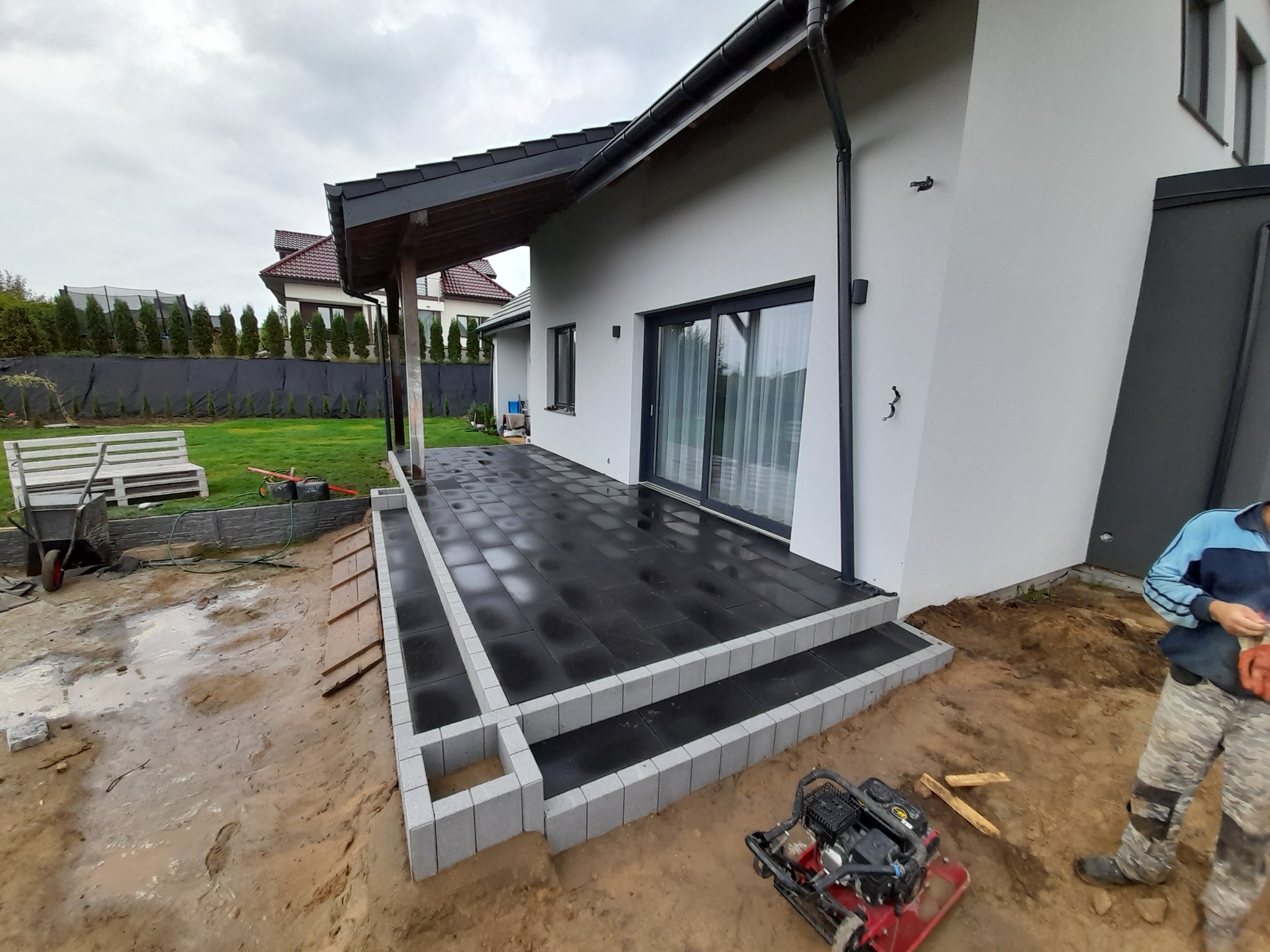 Nowoczesny taras z czarnej kostki brukowej z szarymi obrzeżami, widoczny w trakcie budowy, z ubijarką mechaniczną i pracownikiem w tle.