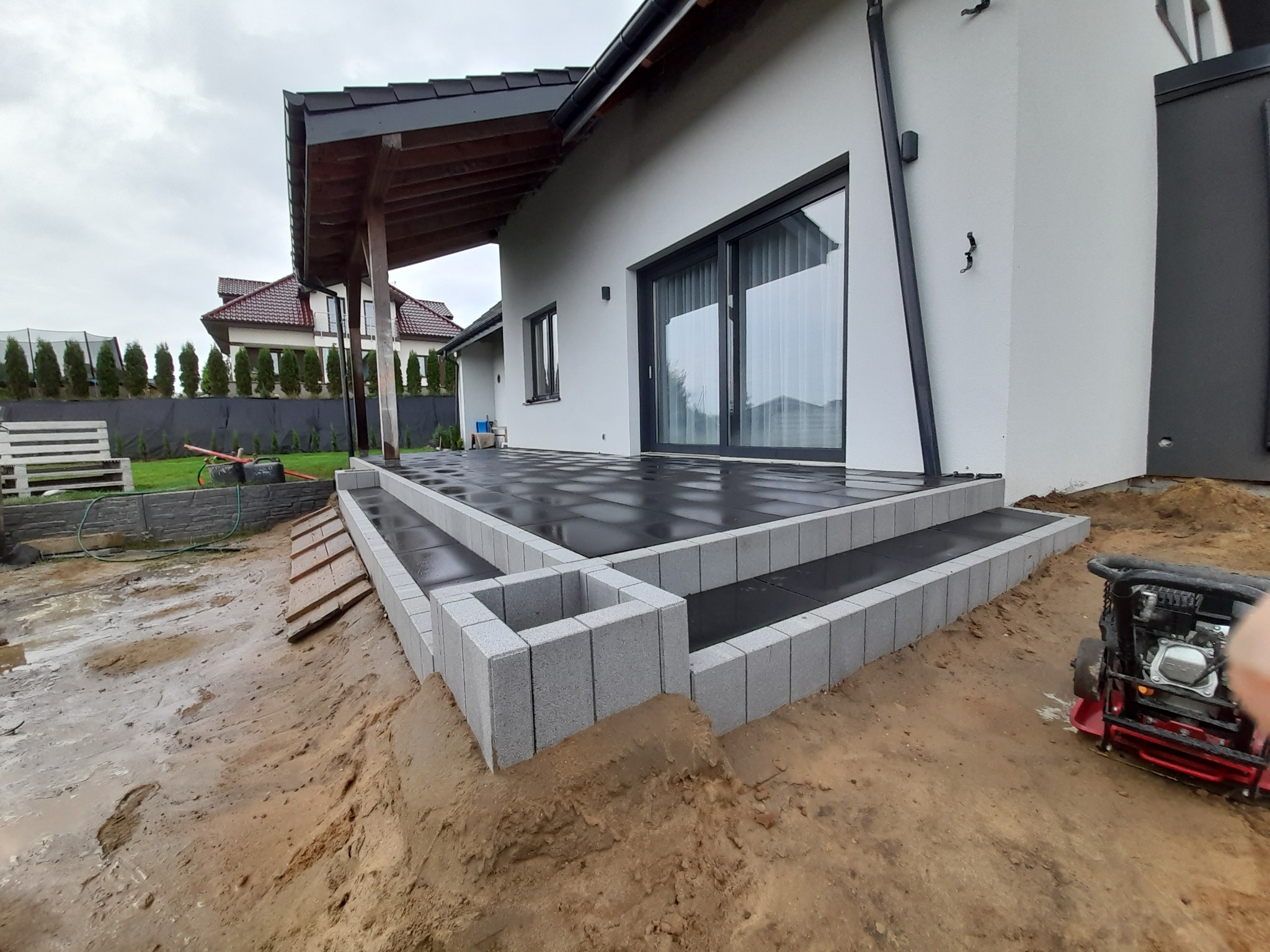 Nowoczesny taras z czarnej kostki brukowej i szarych betonowych stopni, widoczna ubijarka w tle, teren wokół jeszcze niezagospodarowany.