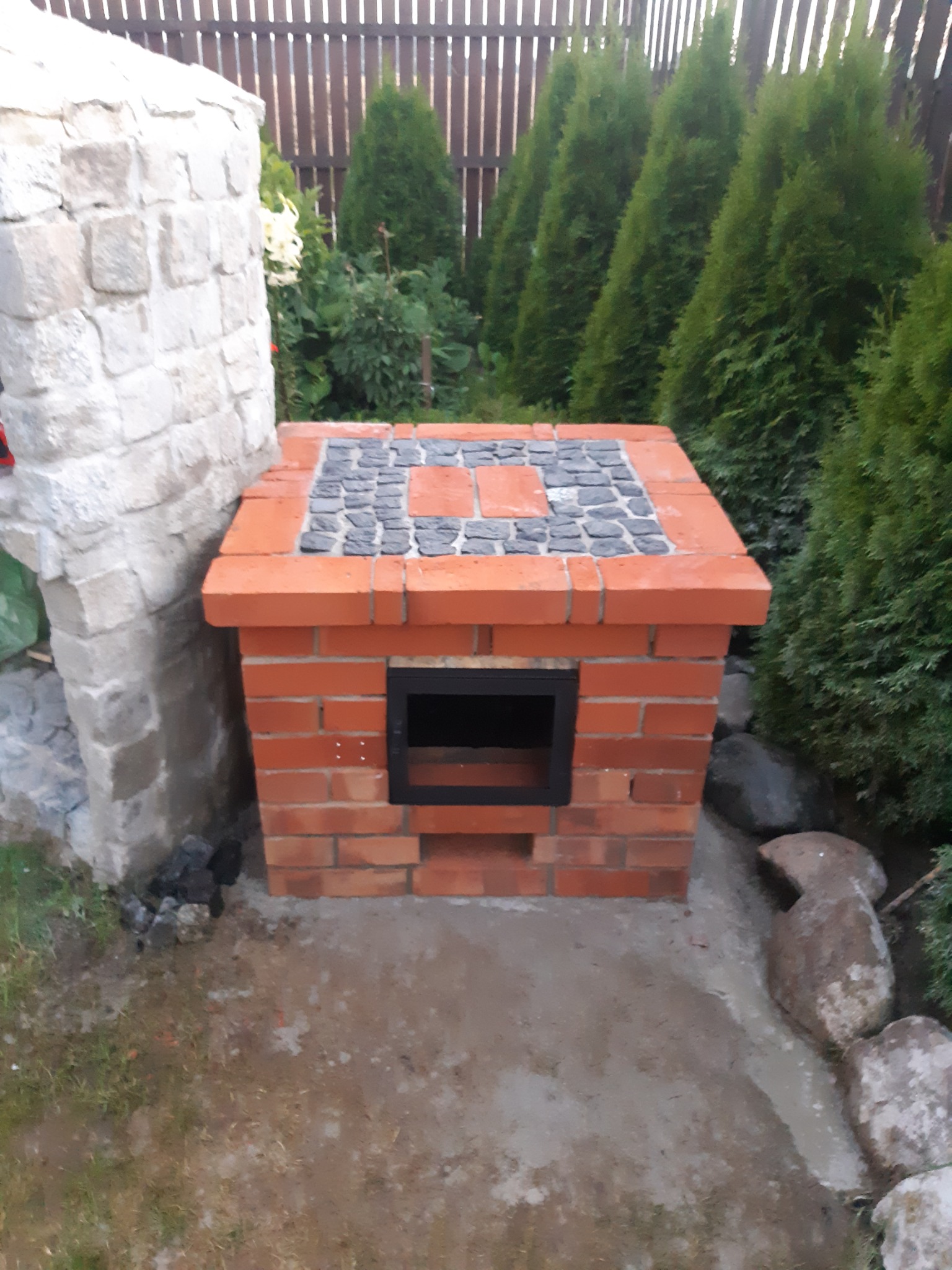 Ogród z murowanym grillem z czerwonej cegły i blatem wykończonym kostką granitową, otoczony tujami i kamieniami.