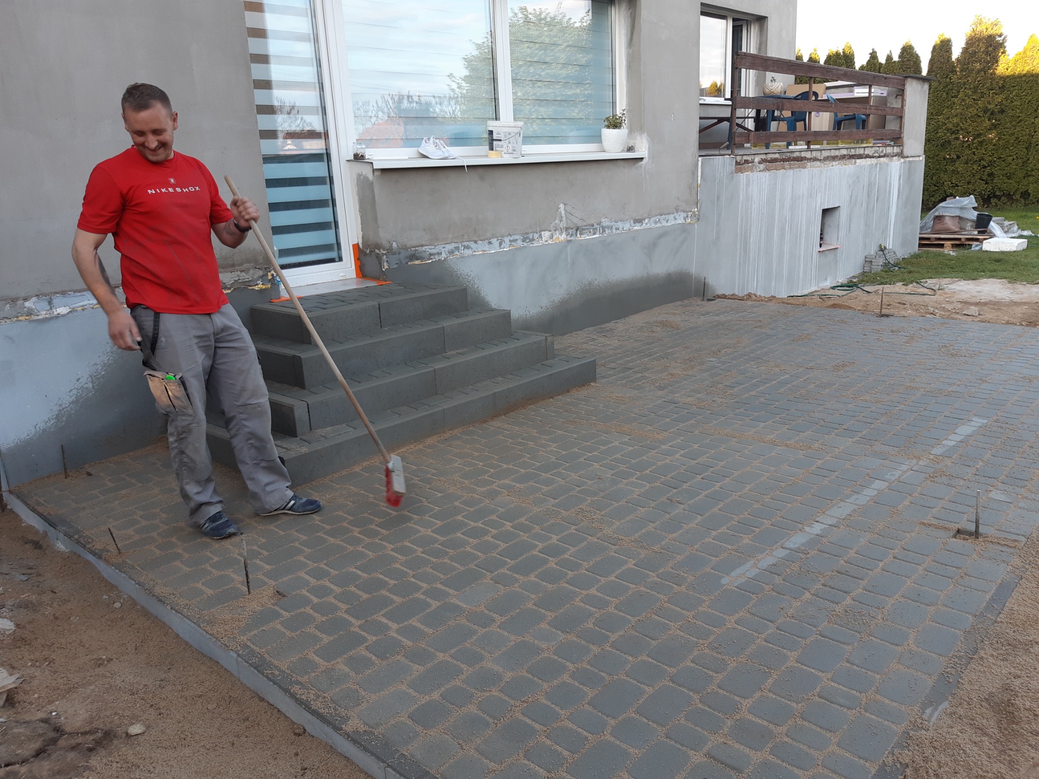 Pracownik zamiata piasek ze świeżo ułożonej, szarej kostki brukowej na podjeździe przed domem z widocznymi schodami wejściowymi i oknem.