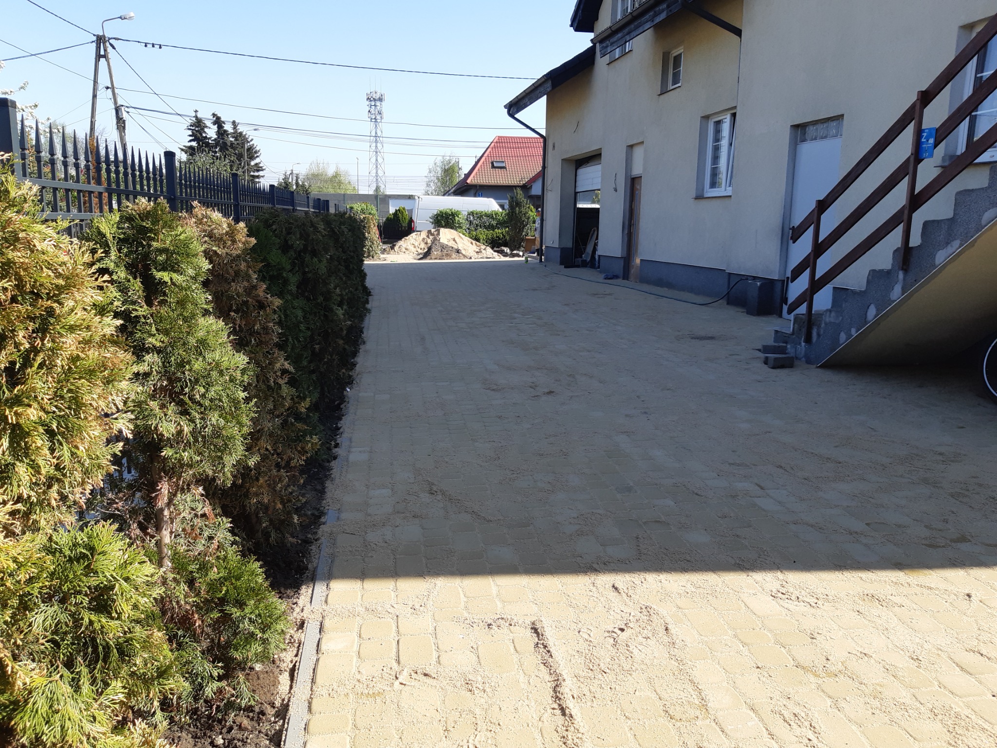 Żółta kostka brukowa ułożona jako podjazd do domu, częściowo zasypana piaskiem, obok żywopłot z tui.