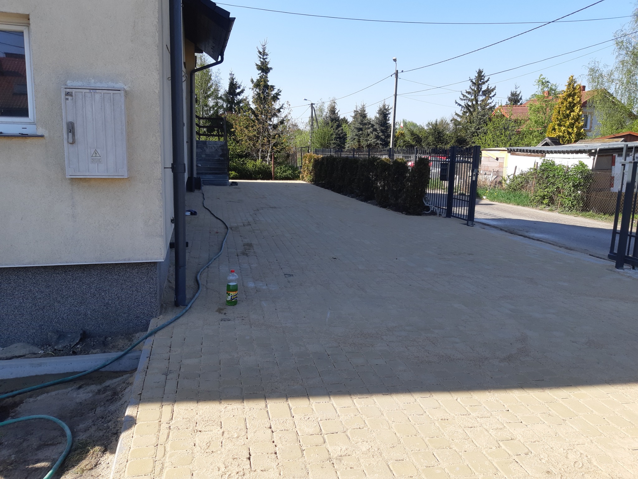 Nowo ułożona, beżowa kostka brukowa na podjeździe do domu jednorodzinnego, widoczny fragment elewacji budynku, wąż ogrodowy i butelka z płynem na kostce.