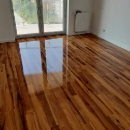 Błyszcząca, nowo ułożona podłoga z paneli drewnopodobnych w pustym pokoju z dużym oknem balkonowym, widoczne odbicie okna w podłodze.