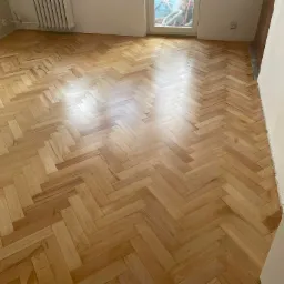 Jasna, odnowiona podłoga parkietowa ułożona we wzór jodełki, widoczna w słonecznym pokoju z grzejnikiem i oknem w tle.